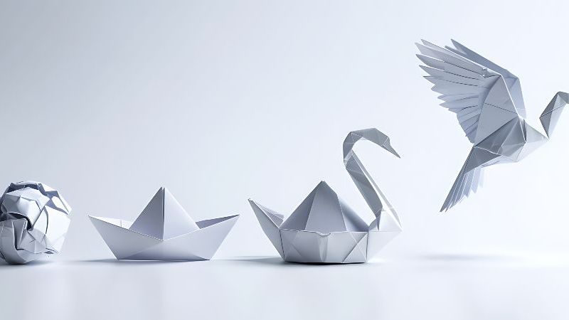 Das Bild zeigt einen zerknüddelten Papierball, ein gefaltetes Papierschiff, einen Origami-Schwan und einen Origami-Kranich, der in die Luft abhebt.