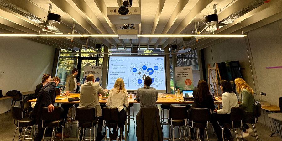 © Battery Incubator Das Bild zeigt Menschen in einem Veranstaltungsraum, die an Tischen sitzen und auf eine Präsentation auf einer Leinwand schauen.