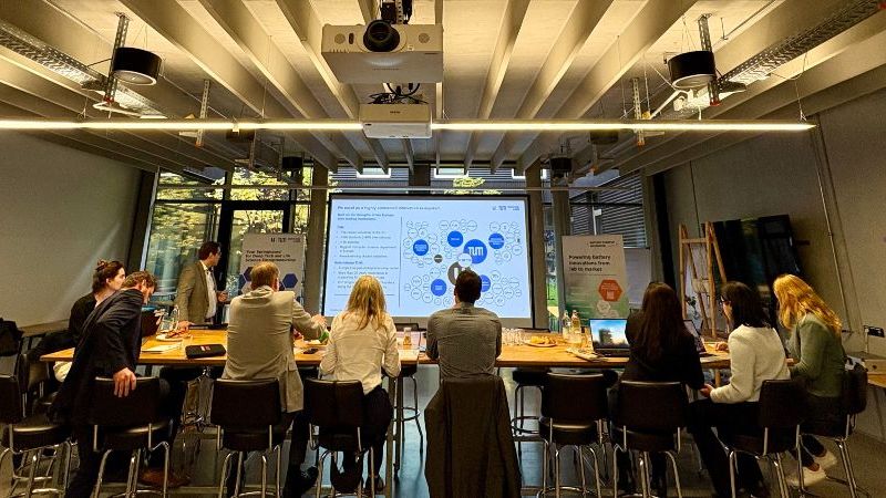 © Battery Incubator Das Bild zeigt Menschen in einem Veranstaltungsraum, die an Tischen sitzen und auf eine Präsentation auf einer Leinwand schauen.