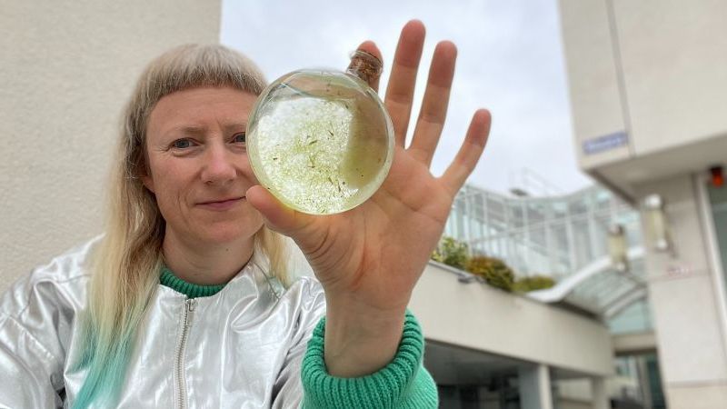 Das Bild zeigt Line Krom auf einer Ausstellung im Nordwestzentrum in Frankfurt. Sie hält ein Glasgefäß in der Hand, in dem sich Algen im Wasser befinden.