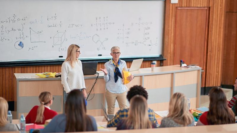 Das Bild zeigt eine Professorin mit einer Kollegin in einem Hörsaal vor einer mit mathematischen Formeln beschriebenen Tafel.