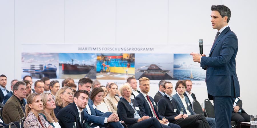 Das Bild zeigt einen Referenten auf einer Bühne; im Hintergrund befindet sich ein großes Poster mit Fotos aus dem maritimen Bereich..