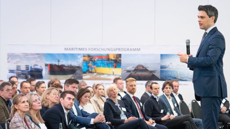 Das Bild zeigt einen Referenten auf einer Bühne; im Hintergrund befindet sich ein großes Poster mit Fotos aus dem maritimen Bereich..