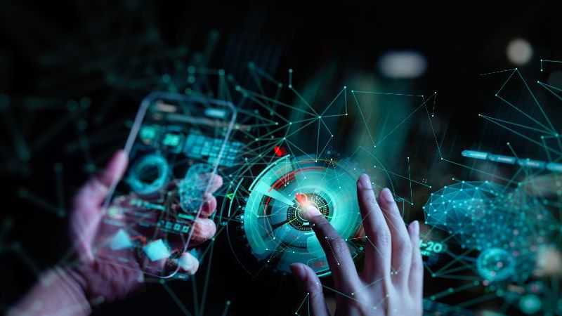 Das Bild zeigt eine futuristische digitale Benutzeroberfläche mit einer Hand am Smartphone und einer weiteren Hand, die interaktive holografische Elemente, Netzwerke und Datenvisualisierungen in einer dunklen Hightech-Umgebung steuert.