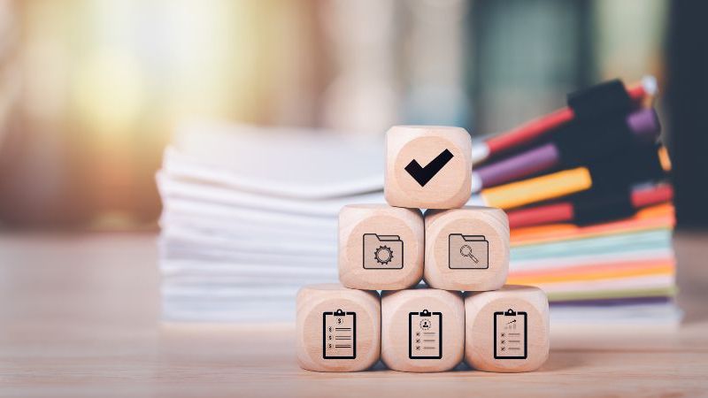 © chaylek – stock.adobe.com Das Bild zeigt verschiedene Holzwürfel mit Symbolen aus dem Bereich des Monitorings und der Evaluation; im Hintergrund liegt ein Stapel mit Akten..