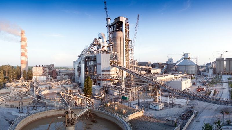 © dmitrykobets – stock.adobe.com Das Bild zeigt eine großflächige Industrieanlage mit Produktionsgebäuden, Silos, Förderanlagen, Rohrleitungen und einem Schornstein.