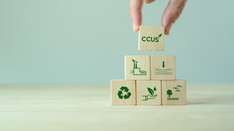 Das Bild zeigt eine Hand, die einen Holzwürfel mit der Aufschrift „CCUS“ auf weitere Holzwürfel mit grünen Symbolen für Klimaschutz, Nachhaltigkeit, Recycling und industrielle CO₂-Speicherung setzt.
