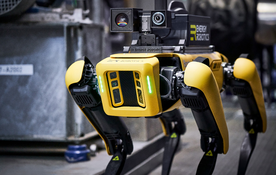 Ein gelb-schwarzer Industrieroboter auf vier Beinen in einem Raum voller metallener Behälter und Rohrleitungen.