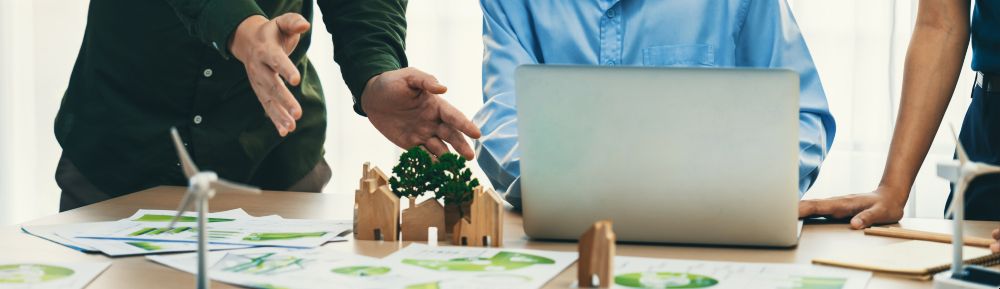 Drei Menschen sitzen an einem Tisch und schauen auf einen Laptop. Auf dem Tisch liegen mehrere Pläne. Außerdem ist aus Modell-Häusern, Modell-Windkraftanlagen und Modell-Bäumen eine Landschaft erbaut.