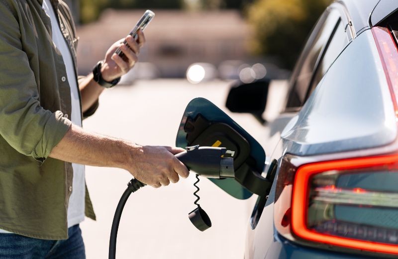 Auf dem Bild ist ein Elektroauto zu sehen, das von einer Person für den Ladevorgang vorbereitet wird. Die Person hat außerdem ein Handy in der Hand.