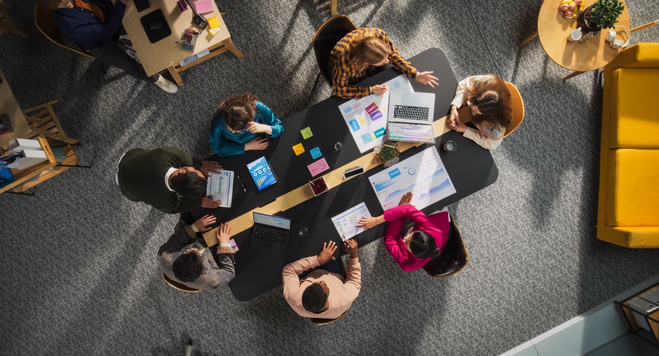 Auf dem Bild sieht man einen modernen Besprechungsraum. An einem Tisch sitzen sechs junge Leute, eine Person steht daneben.