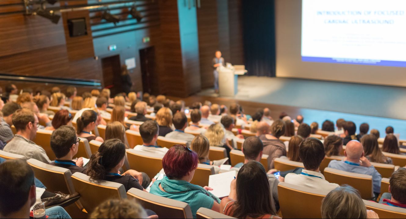 Auf dem Bild ist ein Hörsaal voller Studenten und einem Professor zu sehen.
