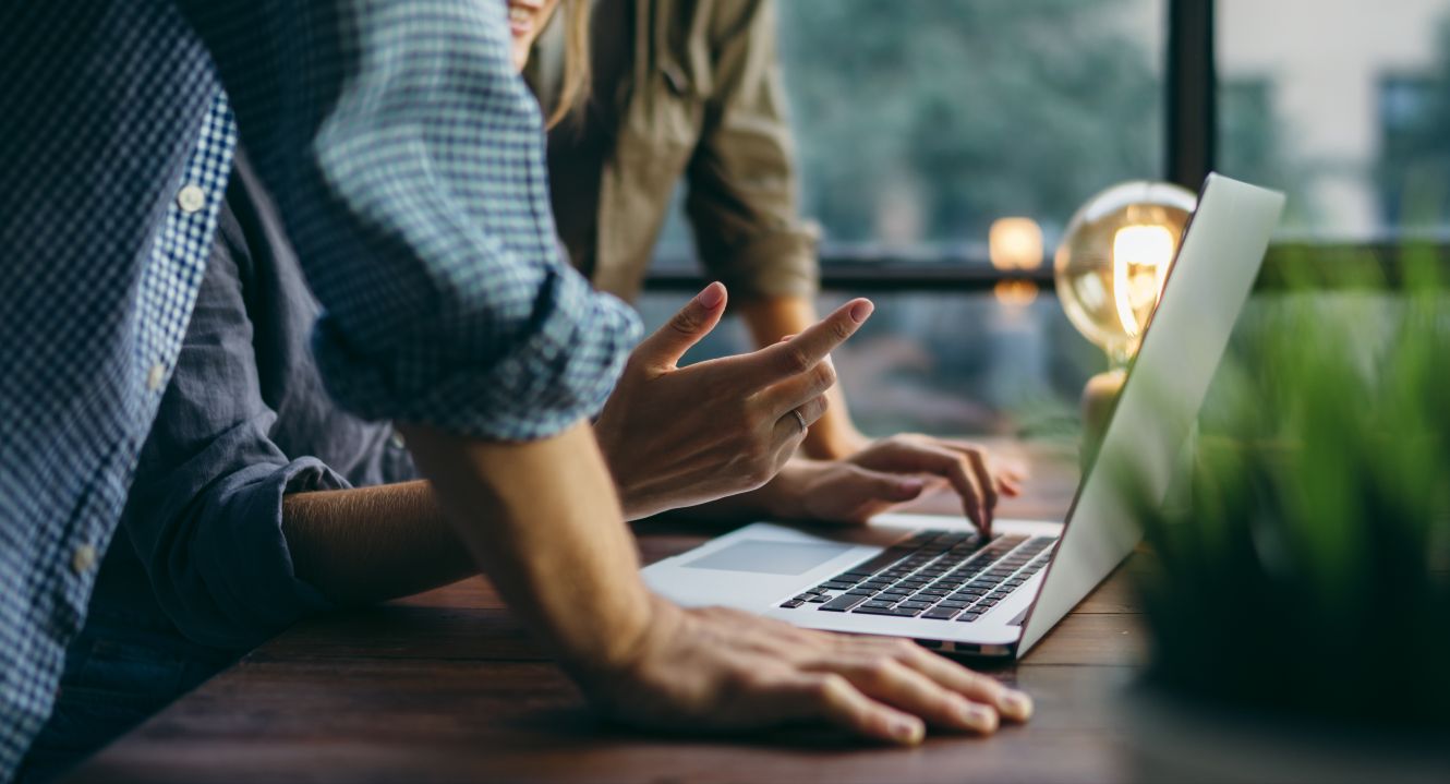 Auf dem Bild sieht man zwei Personen, die über ein Laptop gebeugt sind. Daneben liegt eine Glühbirne.