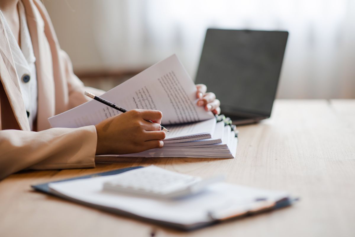 A person sits at a table and writes notes on a stack of paper. A laptop is in the background.