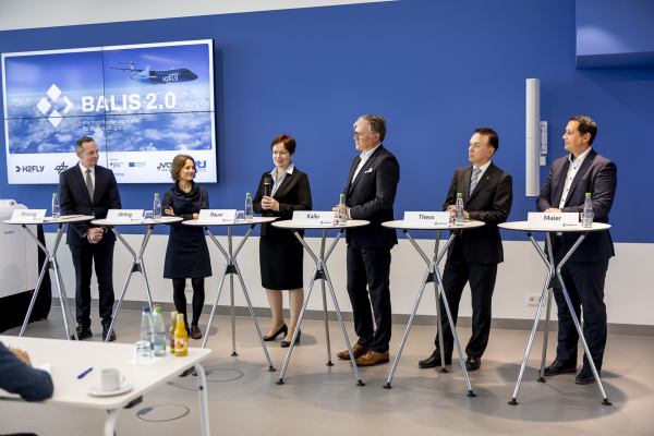 Das Bild zeigt die PtJ-Leitung Dr. Stefanie Bauer hinter einem Podium in ein Mikrofon sprechend, umgeben von weiteren Personen. 