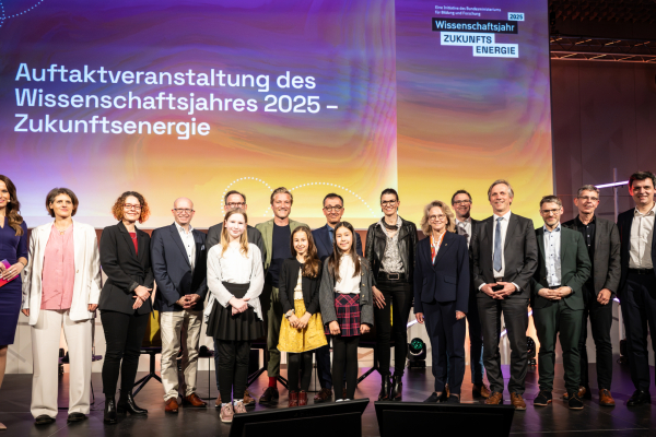 Das Bild zeigt Menschen auf einer Bühne. Im Hintergrund ist der Schriftzug 