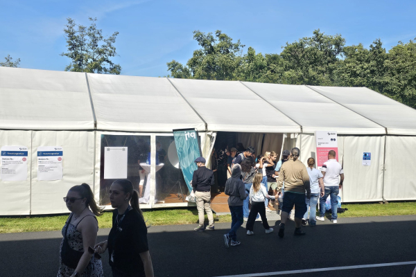 Besucher:innen stehen vor unserem weißen Ausstellungszelt; einige treten gerade in den Stand ein, andere schlendern an der sonnigen Außenseite vorbei. 