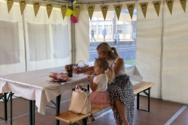 Eine Frau sitzt mit einem kleinen Mädchen an einem Basteltisch in unserem Ausstellungszelt; vor ihnen liegen Buntstifte und Papier, über ihnen hängen gelbe PtJ-Wimpel. 