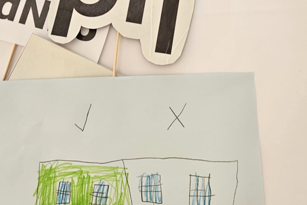 Kinderzeichnung auf einem Blatt Papier; darüber liegen Bastelmaterialien und PtJ-Schilder. 