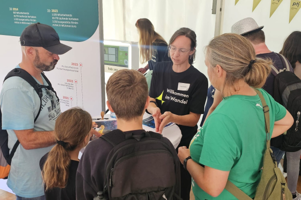 Eine PtJ-Mitarbeiterin erklärt Besucher:innen an einem Informationsstand Inhalte; um sie herum stehen Erwachsene und Kinder, die aufmerksam zuhören. 