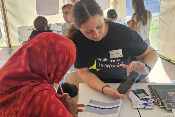 Eine PtJ-Mitarbeiterin im schwarzen T-Shirt mit der Aufschrift ‚Willkommen im Wandel‘ erklärt einer Besucherin im roten Kopftuch etwas auf dem Smartphone und zeigt dazu ein Infoblatt; beide sitzen an einem Tisch im Zelt. 