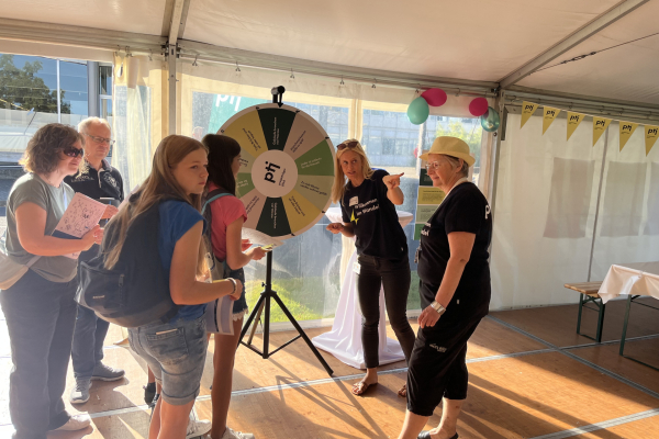 Kinder stehen fröhlich vor einem Glücksrad-Stand („PtJ“), an dem eine Mitarbeiterin mit Strohhut sie betreut; im Hintergrund ein heller Pavillon mit sonniger Atmosphäre. 
